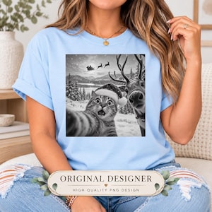 May include: A light blue t-shirt featuring a graphic print of a cat wearing a Santa hat taking a selfie with a reindeer in a snowy landscape. In the background, a sleigh with reindeer flies across the sky above a forest of pine trees.
