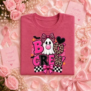 May include: A pink t-shirt featuring a graphic of a white ghost with a pink bubblegum bubble, a leopard print bow, and the text BOO CREW in pink and leopard print. The design includes black bats, stars, a black heart, and a checkered pattern.