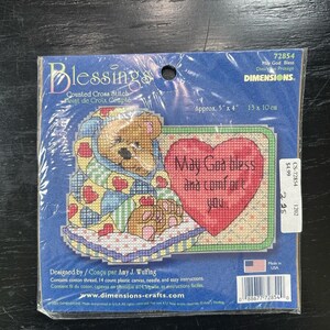 Op de afbeelding: Een kruissteekpakket met een teddybeer, gewikkeld in een deken met hart- en stermotieven. De set bevat de tekst "May God bless and comfort you" op een hartvormig ontwerp. Geschatte afmetingen: 13 x 10 cm.