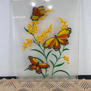Puede incluir: Panel decorativo rectangular transparente con tres mariposas de alas naranjas y amarillas. Las mariposas están colocadas alrededor de acentos florales amarillos y tallos verdes. El panel es transparente y tiene una superficie ligeramente texturizada.
