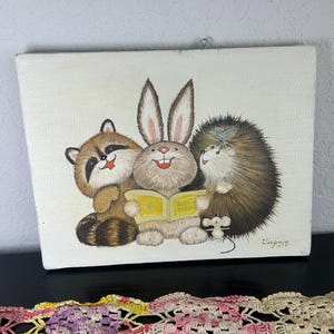 Puede incluir: Pintura vintage que representa un mapache, un conejo y un erizo leyendo un libro. El libro está abierto, con el texto "Baby Book" visible. La obra está firmada "Virginia 1979". La pintura está sobre un lienzo blanco, con un lazo para colgar.