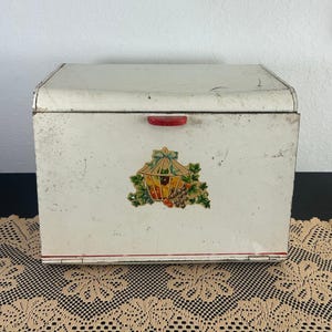 Puede incluir: Panera vintage blanquecina con asa roja y diseño decorativo de frutas y linterna. La caja rectangular tiene una parte superior curvada y una franja roja a lo largo del borde inferior. La caja es de metal.