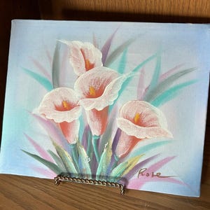 Puede incluir: Pintura original de calas en tonos rosa, naranja y blanco, con hojas verdes y moradas. La obra está sobre lienzo, expuesta sobre un soporte metálico decorativo. La firma del artista, "Rose", es visible. Medidas aproximadas: 20 cm x 25 cm.