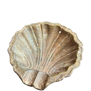 May include: A decorative, antique-style seashell-shaped dish. The dish is made of a bronze-colored metal with a textured surface. The shell design features detailed ridges and a scalloped edge. The dish is likely used for holding small items.