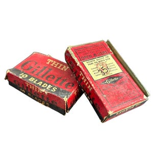 Puede incluir: Dos cajas vintage rojas y negras de cuchillas Gillette Thin 10. Una caja está abierta, revelando las cuchillas. Las cajas tienen texto que dice "Gillette" y "Thin" en letras blancas. Las cajas están ligeramente desgastadas.
