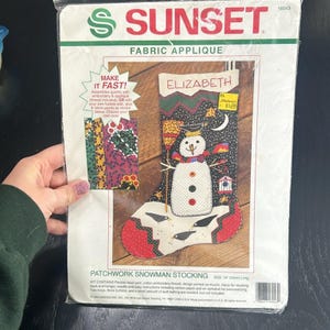 May include: A craft kit for making a patchwork snowman stocking. The kit includes fabric, yarn, embroidery thread, and instructions. The stocking is 16 inches long and features a snowman with a black hat and a red and white striped scarf. The stocking is decorated with black birds and a moon.