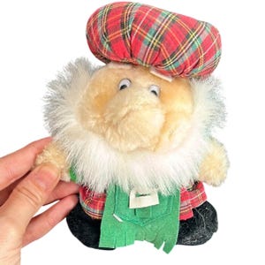May include: A soft toy doll featuring a cream-coloured face, white fur trim, and a red plaid hat. The doll is dressed in a red plaid shirt, green overalls, and black shoes. The doll is held by a hand.