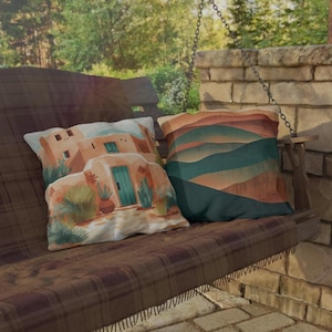 May include: Two decorative throw pillows on a wooden swing. One pillow features a Southwestern-style building scene with a turquoise door. The other pillow has a landscape design with layered earth-tone colors.