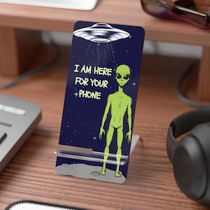 May include: A phone stand featuring a green alien under a UFO, with the text "I AM HERE FOR YOUR PHONE" on a dark blue background. The stand is made of clear acrylic and is designed to hold a phone.