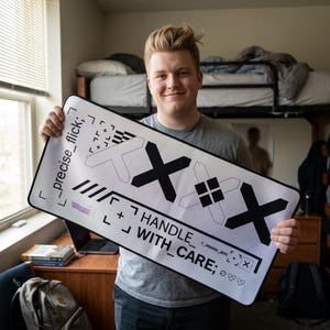 May include: A rectangular white desk mat featuring black graphic designs, including large X shapes, geometric patterns, and the text "precise_flick;" and "HANDLE_WITH_CARE;" along with "<steady_aim;" and small heart symbols.