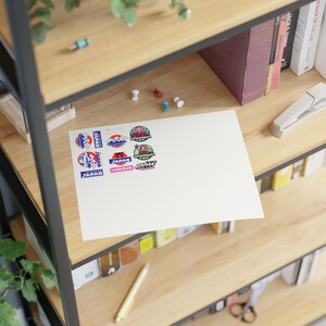 Puede incluir: Una hoja rectangular blanca que presenta una colección de pegatinas coloridas con varios diseños, incluyendo textos como Discover Japan, Japan Horizon y Super Wheelspin, dispuestos en dos columnas sobre la superficie del papel.