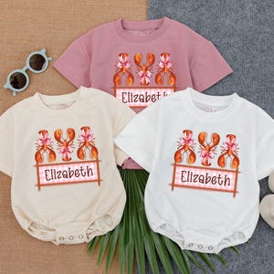 May include: Three baby onesies with a lobster graphic and the name "Elizabeth" printed on each. The onesies are in white, cream, and pink.