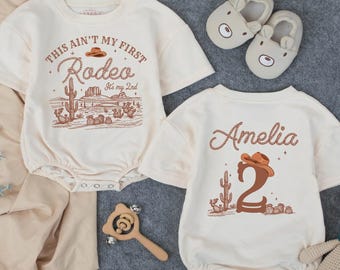 May include: Two cream-colored baby rompers with cowboy-themed graphics. One says "This ain't my first Rodeo" with a desert scene. The other has the name "Amelia" and a number 2. Also, a pair of bear-shaped slippers and a teddy bear.