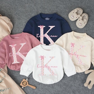 May include: Four children's sweatshirts in navy, pink, white, and cream. Each features a floral letter "K" and the name "Kinsley mae" in pink script. Accessories include bear slippers, a wooden toy, and a stuffed bear.