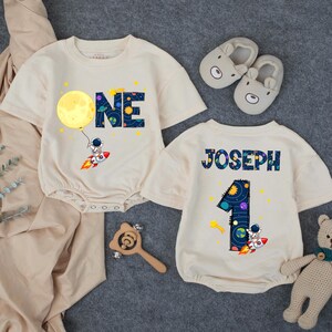May include: Two cream-colored baby onesies with a space theme. One has the word "ONE" with a moon and astronaut graphic. The other has the name "JOSEPH" and the number "1" with a similar space design. Also included are bear slippers, a wooden rattle, and a teddy bear.