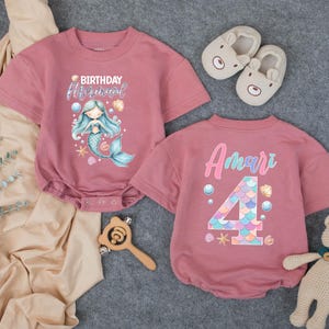 May include: Two dusty rose-colored baby bodysuits. One features a mermaid graphic with the text "Birthday Mermaid". The other has the name "Amari" and the number "4" with a mermaid scale design. Accessories include bear slippers and a stuffed bear.