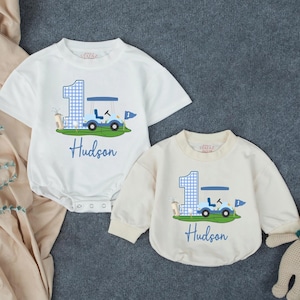 May include: Two white baby bodysuits featuring a blue gingham number one, a golf cart, and the name Hudson printed in blue. The design includes a golf bag and flag on a green patch. One bodysuit has short sleeves, and the other has long sleeves.