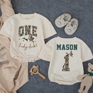 Puede incluir: Dos peleles de beb&eacute; color crema con mangas cortas. Uno dice "ONE lucky duck!" con un gr&aacute;fico de pato y letras de camuflaje. El otro dice "MASON" y tiene un gr&aacute;fico de pato y el n&uacute;mero "1" en camuflaje. Tambi&eacute;n hay un par de zapatillas de oso y un osito de peluche.