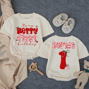 May include: Two cream-colored baby rompers with red strawberry and text designs. One says "It's my Berry First Birthday" and the other says "Sophie 1". The image includes a pair of gray bear slippers, a wooden rattle, and a stuffed bear.