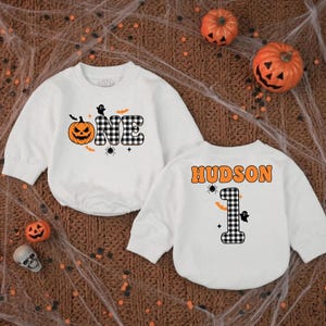 May include: Two white long-sleeve sweatshirts featuring Halloween graphics. One sweatshirt displays the word ONE with a pumpkin and black and white plaid letters. The other sweatshirt features the name HUDSON above a plaid number 1.