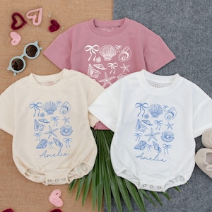 May include: Three baby outfits: a light pink t-shirt, a cream-colored romper, and a white romper. Each garment features a blue seashell and bow design. The cream romper has the name "Amelia" printed in blue. Sunglasses and baby shoes are also visible.