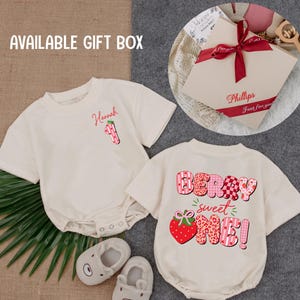May include: Cream-colored baby rompers with strawberry-themed graphics and text. One says "Hannah 1" and the other says "Berry Sweet One!". Includes a gift box with a red ribbon and a pair of gray bear-shaped baby shoes.