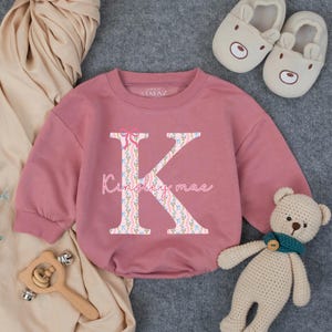 May include: A dusty rose baby onesie with long sleeves. The onesie features a large floral letter "K" with a pink bow and the name "Kinsley mae" in script. A pair of bear slippers, a wooden toy, and a teddy bear are also in the image.