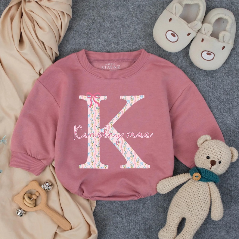 May include: A dusty rose baby onesie with long sleeves. The onesie features a large floral letter "K" with a pink bow and the name "Kinsley mae" in script. A pair of bear slippers, a wooden toy, and a teddy bear are also in the image.
