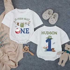 May include: Two white short-sleeved baby bodysuits. The left one features a fishing graphic with the text O-FISH-ALLY ONE and a red and white bobber. The right one displays the name HUDSON above a large blue number 1 with fishing gear.