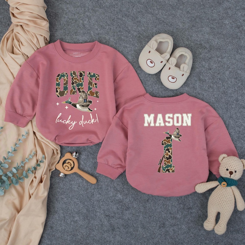 Puede incluir: Dos sudaderas rosas para beb&eacute;s. Una tiene "ONE" en estampado de camuflaje con un gr&aacute;fico de pato volando y las palabras "lucky duck!". La otra tiene el nombre "MASON" y un "1" de camuflaje con un gr&aacute;fico de pato.