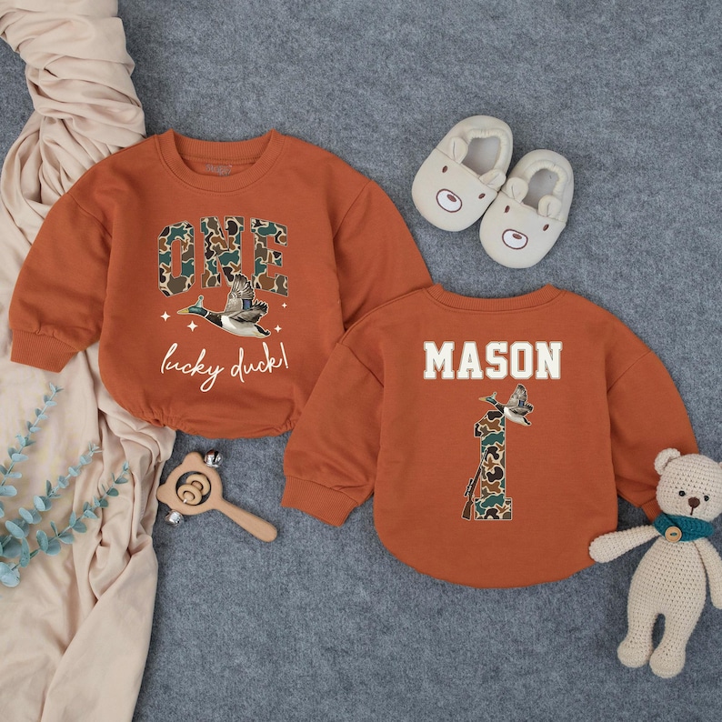Puede incluir: Dos sudaderas para beb&eacute; de color &oacute;xido. Una dice "ONE" en estampado de camuflaje con un gr&aacute;fico de pato y el texto "lucky duck!". La otra dice "MASON" con un "1" de camuflaje y un gr&aacute;fico de pato. Incluye zapatillas de oso y un juguete de madera.