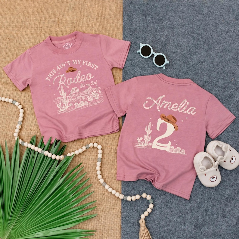 May include: Two pink t-shirts with cowboy-themed graphics. One says "This ain't my first rodeo" and the other has the name "Amelia" and the number "2". Accessories include sunglasses and baby shoes.