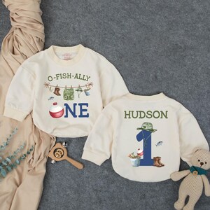 May include: Two cream-colored long-sleeve sweatshirts. One features the text O-FISH-ALLY ONE with fishing gear illustrations and a bobber. The other displays the name HUDSON above the number 1, decorated with a fishing hat, bucket, and boots.