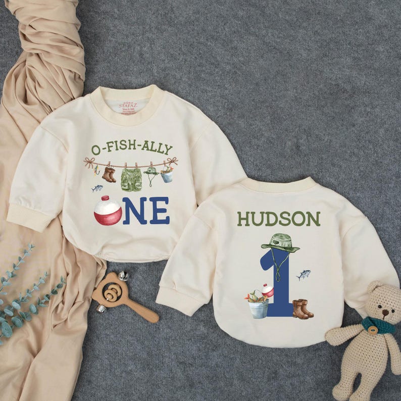 May include: Two cream-colored long-sleeve sweatshirts. One features the text O-FISH-ALLY ONE with fishing gear illustrations and a bobber. The other displays the name HUDSON above the number 1, decorated with a fishing hat, bucket, and boots.