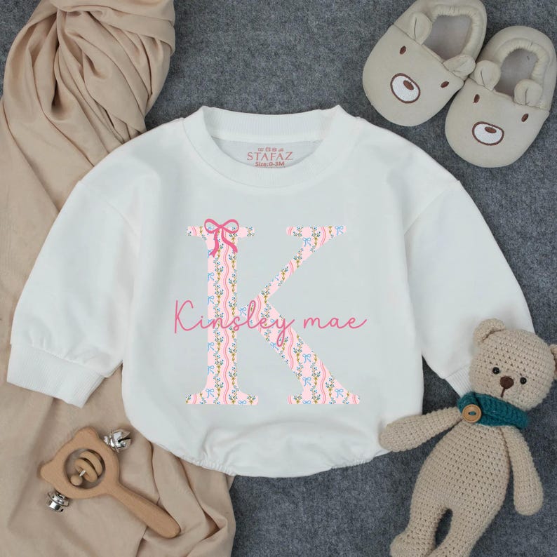 May include: White baby bodysuit with a large pink floral letter "K" and the name "Kinsley mae" in pink script. The bodysuit is surrounded by a teddy bear, baby shoes, and a wooden toy.