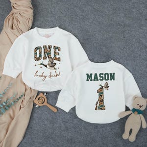 Puede incluir: Dos sudaderas blancas de manga larga. Una presenta la palabra "ONE" en estampado de camuflaje, un gr&aacute;fico de pato volando y el texto "lucky duck!". La otra tiene el nombre "MASON" y un "1" de camuflaje con un gr&aacute;fico de pato.
