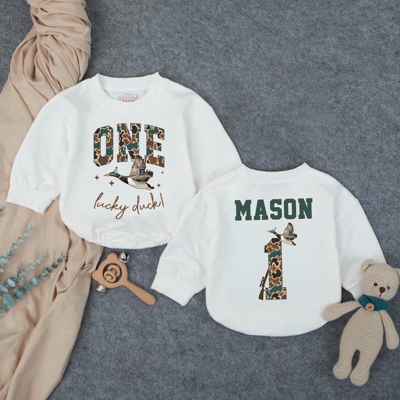 Puede incluir: Dos sudaderas blancas de manga larga. Una presenta la palabra "ONE" en estampado de camuflaje, un gr&aacute;fico de pato volando y el texto "lucky duck!". La otra tiene el nombre "MASON" y un "1" de camuflaje con un gr&aacute;fico de pato.