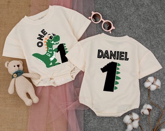 May include: Two cream-colored short-sleeved onesies with a dinosaur theme. One onesie has a green dinosaur with sunglasses holding a black number one. The other onesie has a black number one with green dinosaur scales and the name "DANIEL" in black letters.