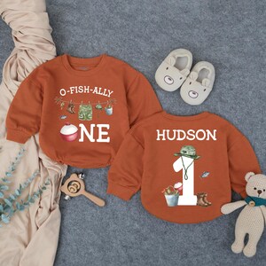 May include: Two rust-colored long-sleeve sweatshirts. One features the text O-FISH-ALLY ONE with fishing gear graphics. The other features the name HUDSON, the number 1, and fishing gear graphics. Includes beige baby shoes and a teddy bear.