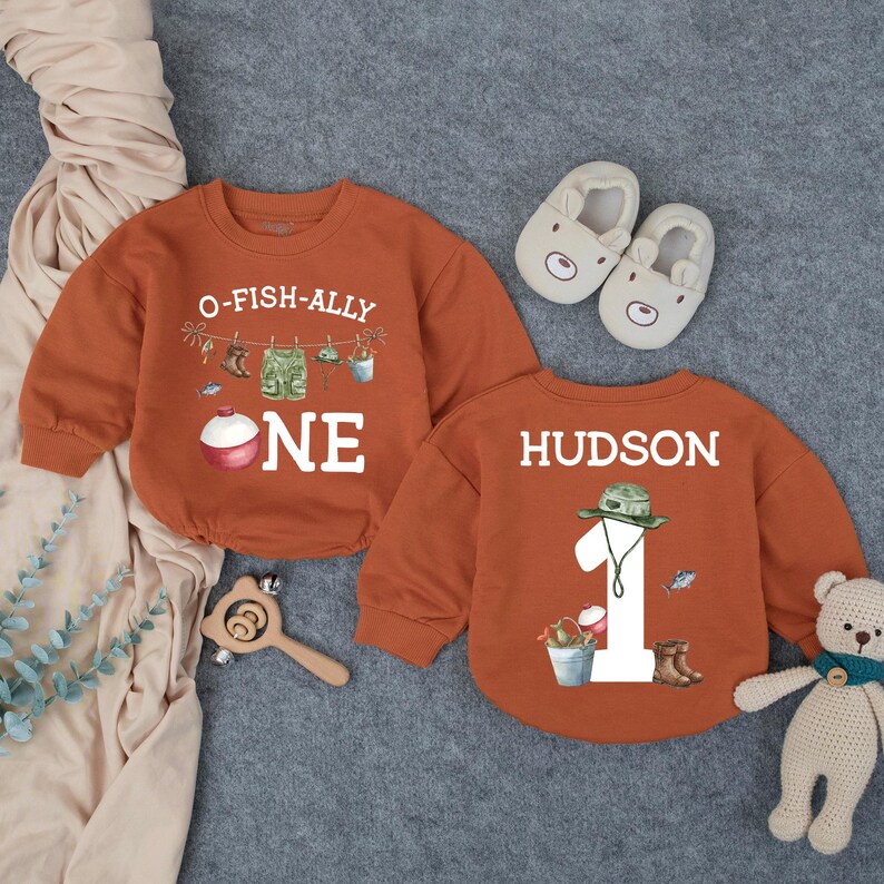 May include: Two rust-colored long-sleeve sweatshirts. One features the text O-FISH-ALLY ONE with fishing gear graphics. The other features the name HUDSON, the number 1, and fishing gear graphics. Includes beige baby shoes and a teddy bear.