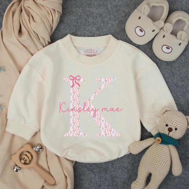 May include: Cream-colored baby romper with a floral "K" initial and the name "Kinsley mae" in pink script. The romper has long sleeves and a round neckline. A pair of bear-shaped slippers, a wooden toy, and a teddy bear are also in the image.