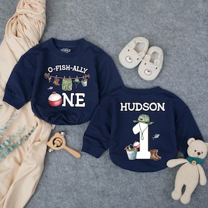 May include: Two navy blue long-sleeve sweatshirts featuring fishing-themed graphics. One shirt displays the text O-FISH-ALLY ONE with a fishing line and bobber. The other shirt features the name HUDSON, the number 1, and fishing gear illustrations.