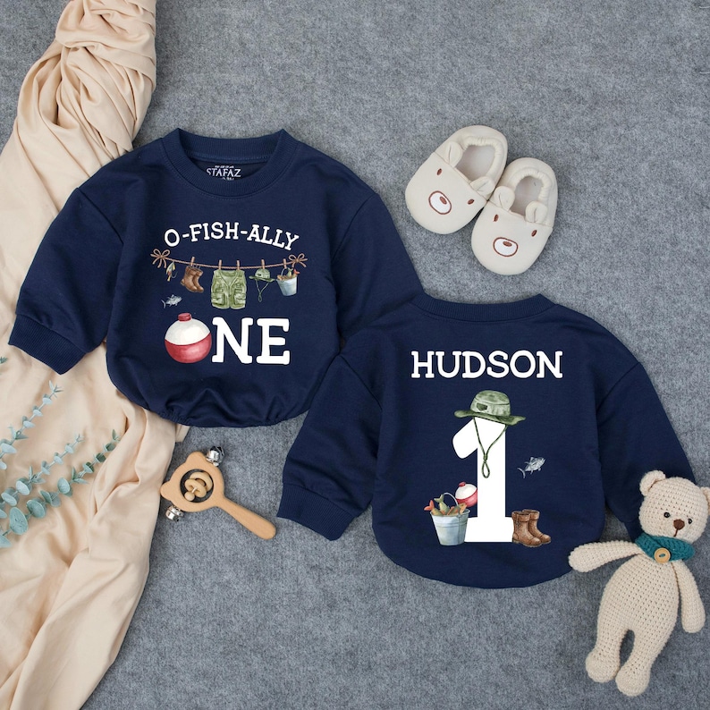 May include: Two navy blue long-sleeve sweatshirts featuring fishing-themed graphics. One shirt displays the text O-FISH-ALLY ONE with a fishing line and bobber. The other shirt features the name HUDSON, the number 1, and fishing gear illustrations.