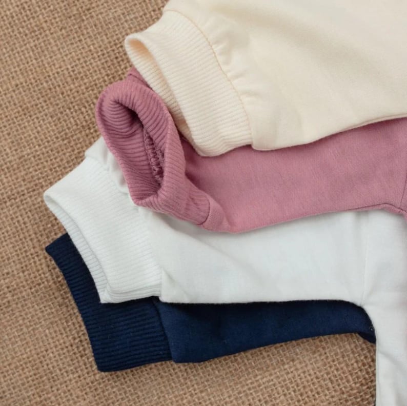 Puede incluir: Primer plano de camisetas de manga larga apiladas en crema, rosa empolvado, blanco y azul marino. Los pu&ntilde;os son acanalados y la tela parece ser de punto suave. Las camisetas est&aacute;n colocadas sobre una superficie texturizada y beige.