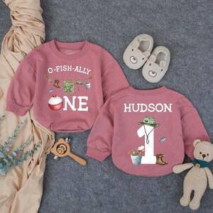 May include: Two dusty rose long-sleeve sweatshirts. One features the text O-FISH-ALLY ONE with fishing gear graphics. The other features the name HUDSON, the number 1, and fishing gear graphics. Includes a pair of beige bear-faced baby shoes.