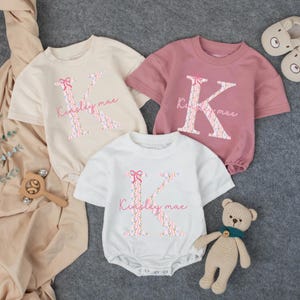 May include: Three baby rompers in cream, pink, and white, each featuring a large floral letter "K" with a pink bow and the name "Kinsley mae". A wooden rattle and a small teddy bear are also visible.