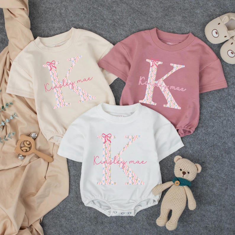 May include: Three baby rompers in cream, pink, and white, each featuring a large floral letter "K" with a pink bow and the name "Kinsley mae". A wooden rattle and a small teddy bear are also visible.