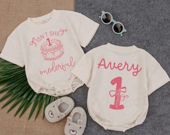 May include: Two cream-colored baby onesies with pink text and graphics. One says "Isn't she onederful" with a cake graphic. The other says "Avery" and a number 1 with a bow. Gray baby shoes and blue sunglasses are also in the image.