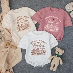 May include: Three baby rompers in cream, white, and dusty rose, each with a short-sleeved design. The front of each romper features a desert-themed graphic with the text "THIS AIN'T MY FIRST Rodeo It's my 2nd". A wooden toy and a teddy bear are also in the image.