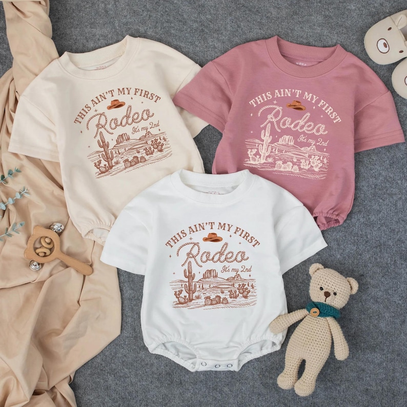 May include: Three baby rompers in cream, white, and dusty rose, each with a short-sleeved design. The front of each romper features a desert-themed graphic with the text "THIS AIN'T MY FIRST Rodeo It's my 2nd". A wooden toy and a teddy bear are also in the image.